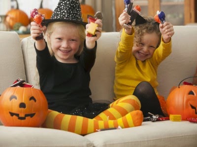 Two girls in Halloween costumes on a couch with jack-o'-lantern buckets, holding up candy