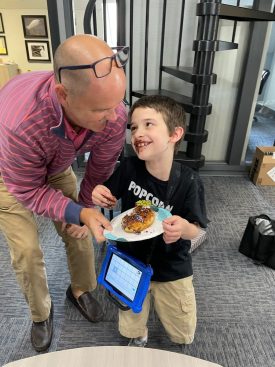 Man helping a smiling boy with a tablet carry a plate of food in an office