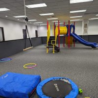 Indoor therapy gym with a colorful play structure and slide, a mini trampoline, a blue mat and hula hoops