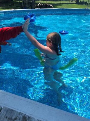 Girl in a pool giving a high five to a person at the pool's edge while holding a pool noodle
